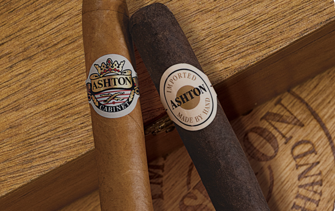 Two Ashton cigars, one light and one dark, resting on a wooden surface, featuring their respective labels.